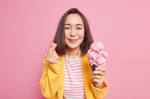 Pleased Asian woman makes mini heart gesture korean like sign holds delicious ice cream eats sweet tasty frozen dessert wears striped t shirt yellow jacket poses against pink wall. Summer time