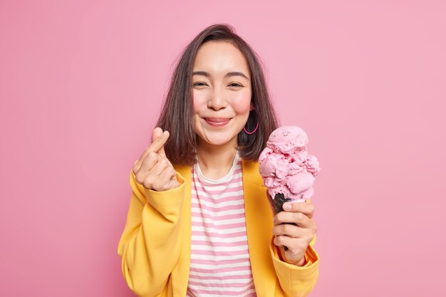 喜んでいるアジアの女性は、看板がおいしいアイスクリームを保持し、甘いおいしい冷凍デザートを食べ、ピンクの壁にストライプのTシャツ黄色のジャケットのポーズをとるようなミニハートジェスチャーを韓国語にします。夏の時間