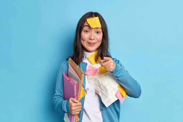 Pleased Asian woman office worker with pleased expression holds a folder and indicates directly at camera