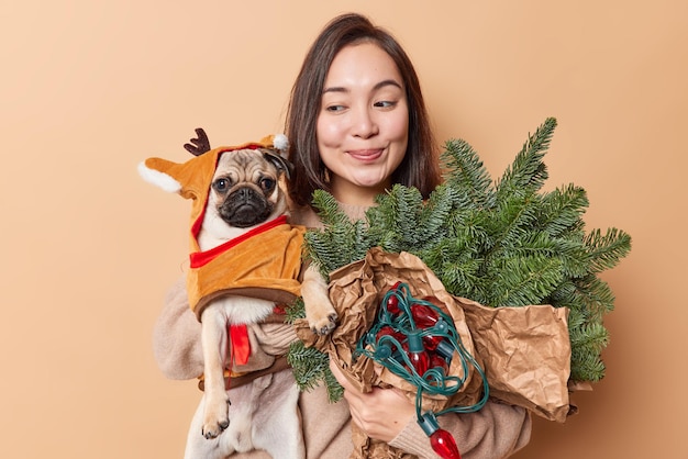 Free photo pleased asian woman prepares for new year poses with pedigree pug dog dressed in winter clothes spruce branches and retro garland for decoration poses against brown background. holidays are coming