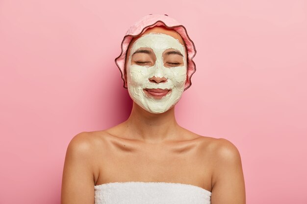 pleased asian woman wears clay purifying organic mask on face, has beauty procedures in spa salon, wears rosy soft protective showercap