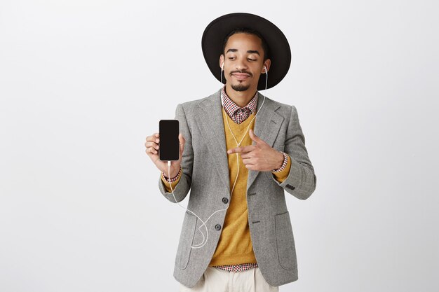 Довольный привлекательный афро-американский мужчина слушает музыку в наушниках и показывает пальцем на экран смартфона, показывая приложение