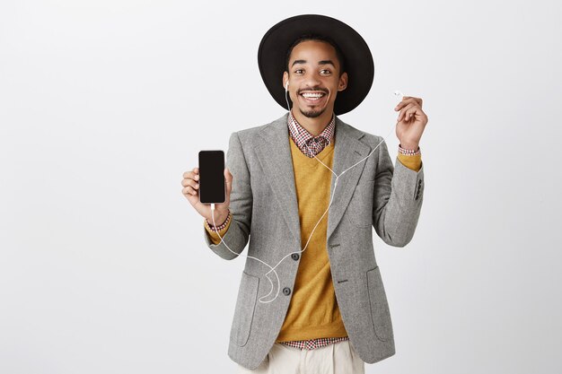 Pleased attractive african-american man listening music in headphones and showing mobile phone screen, application