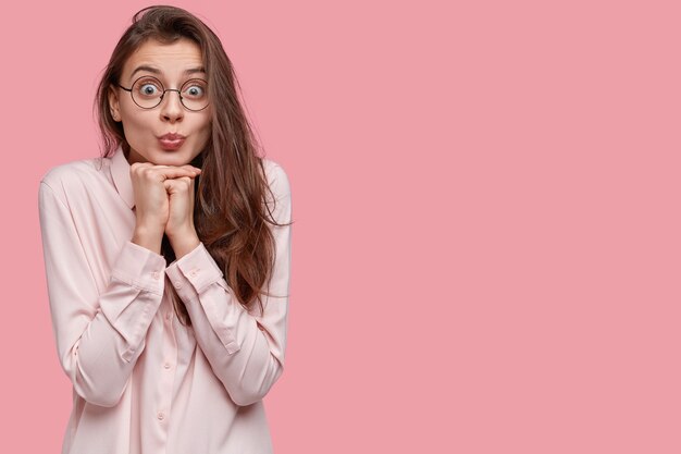 Pleased attractive brunette woman holds hands under chin, dressed in elegant shirt, pouts lips, makes grimace