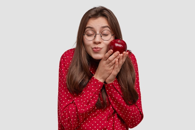 Pleased attractive woman with dark hair, keeps red apple near cheek, closes eyes, has pleasure, wears round glasses