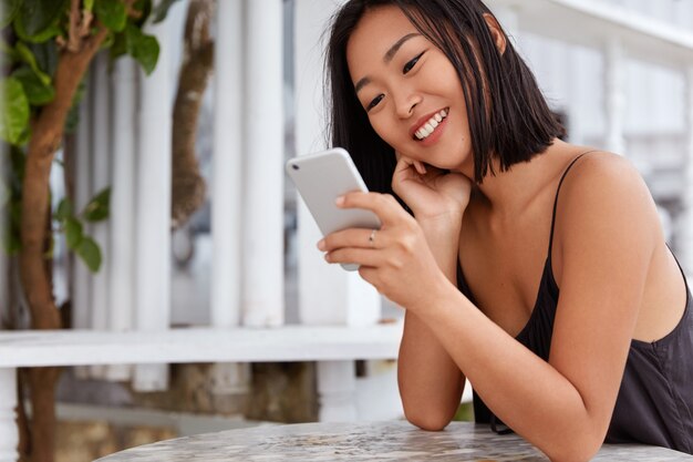 Pleased attractive young Asian woman searches information about summer resort in internet via smart phone, reads good news from website, sits against cafe interior. Woman checks notification