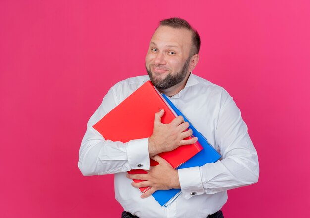 Pleased bearded man wearing white shirt holding folders smiling cheerfully over pink