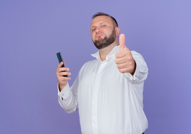 Pleased bearded man wearing white shirt holding smartphone looking  smiling confident showing thumbs up standing over blue wall