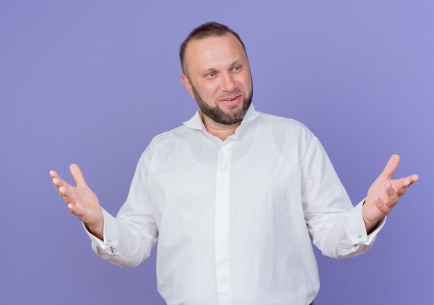 Довольный бородатый мужчина в белой рубашке смотрит в сторону счастливым и позитивным, широко раскинув руки, стоит над синей стеной