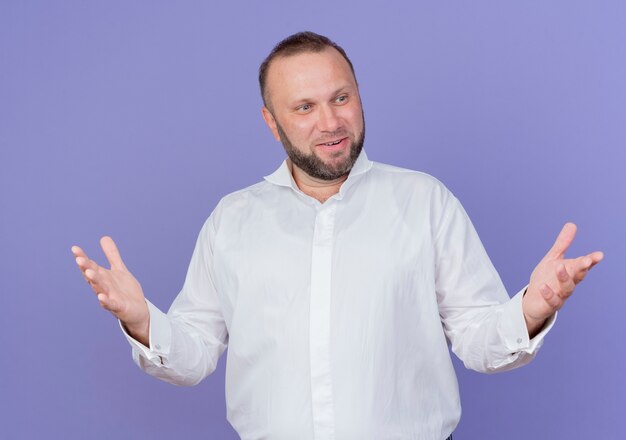 Pleased bearded man wearing white shirt looking aside happy and positive wide spreading arms standing over blue wall