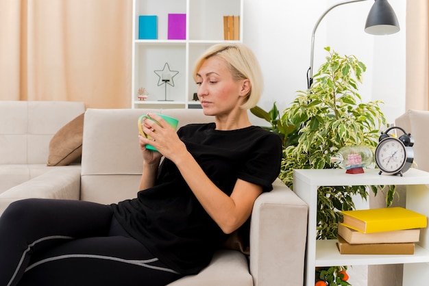 Pleased beautiful blonde russian woman lying on armchair holding and looking at cup inside the living room