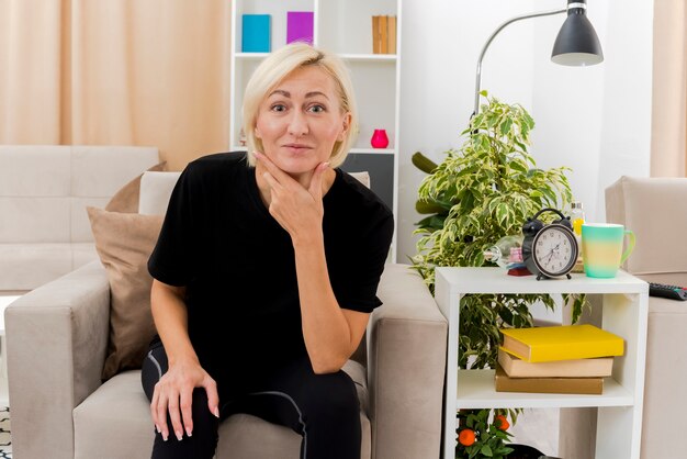 Pleased beautiful blonde russian woman sits on armchair holding chin and looking at camera inside the living room