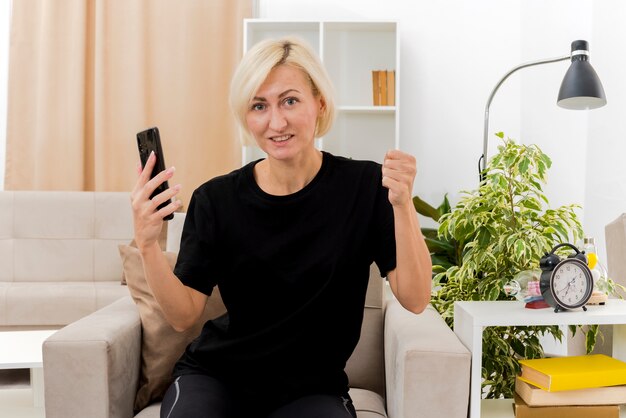 Pleased beautiful blonde russian woman sits on armchair keeping fist and holding phone inside living room