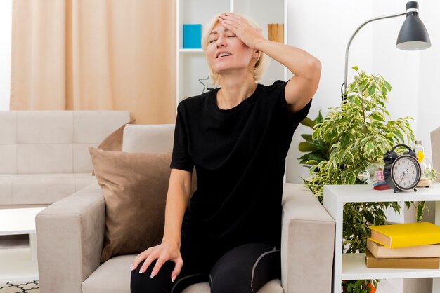 Pleased beautiful blonde russian woman sits on armchair putting hand on forehead inside the living room