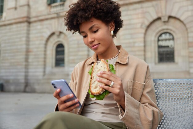 Pleased beautiful curly haired woman eats sandwich and uses mobile phone for surfing internet or chatting online sits on bench against city buildings. People food lifestyle and technology concept