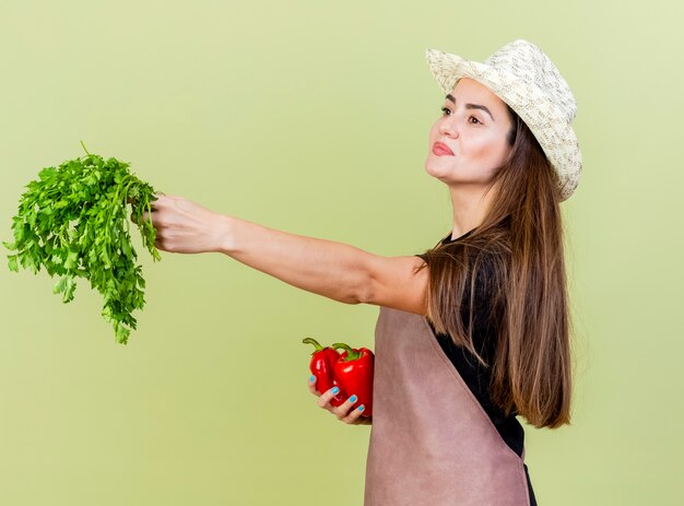 pleased beautiful gardener girl in uniform wearing gardening hat holding peppers and holding out cliantro at side isolated on olive green