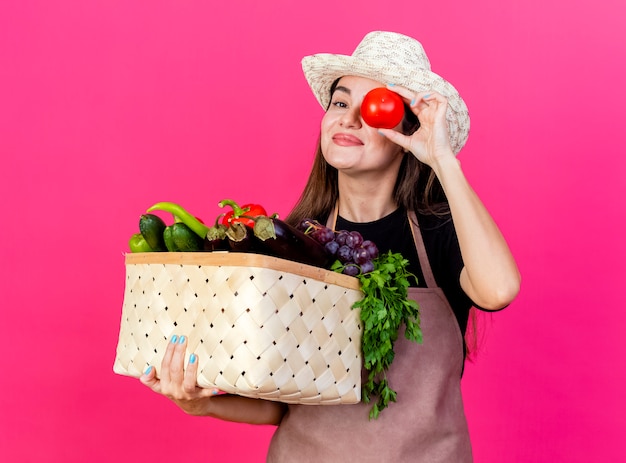 Free photo pleased beautiful gardener girl in uniform wearing gardening hat holding vegetable basket and showing look gesture with tomato isolated on pink background