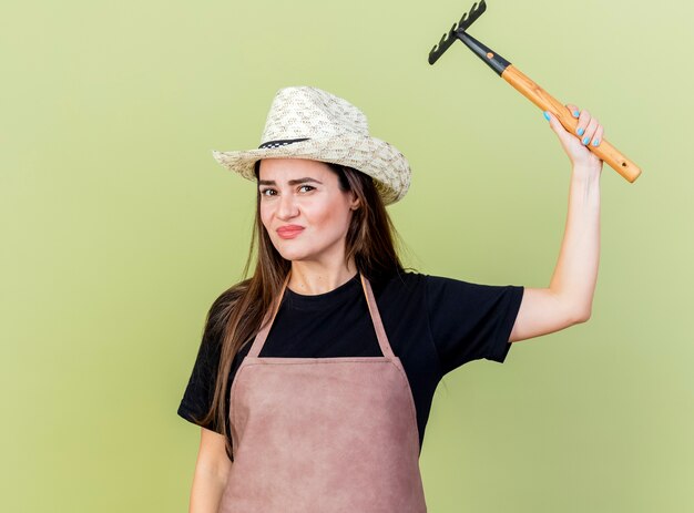 Pleased beautiful gardener girl in uniform wearing gardening hat raising rake isolated on olive green background