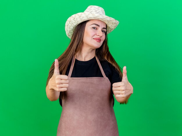 Pleased beautiful gardener girl in uniform wearing gardening hat showing thumbs up isolated on green