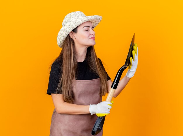 Довольная красивая девушка-садовник в униформе и садовой шляпе с перчатками держит и смотрит на лопату, изолированную на оранжевом фоне