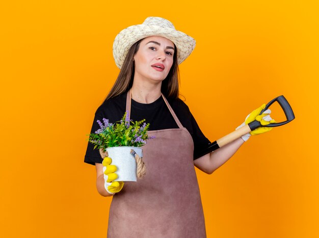肩にスペードを置き、オレンジ色の背景に分離された植木鉢に花を保持している手袋と制服と園芸帽子を身に着けている美しい庭師の女の子を喜ばせる