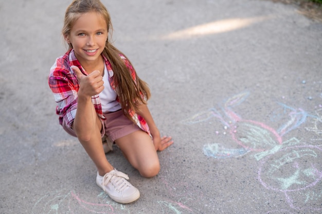 Foto gratuita bella ragazza contenta che approva i suoi disegni di gesso sul terreno