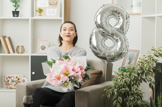 Pleased beautiful girl on happy women day holding bouquet sitting on armchair in living room