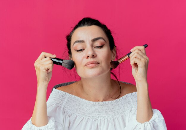 Pleased beautiful girl holds and puts makeup brushes on face isolated on pink wall