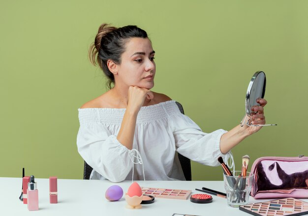 Pleased beautiful girl sits at table with makeup tools puts hand on chin holding and looking at mirror isolated on green wall