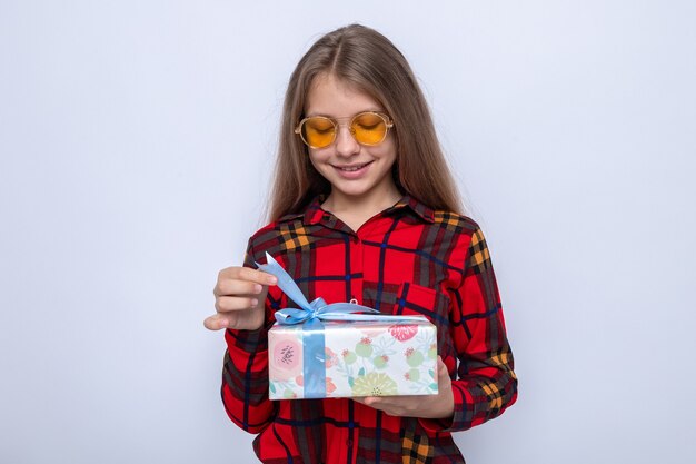 Pleased beautiful little girl wearing red shirt and glasses holding and looking at present 