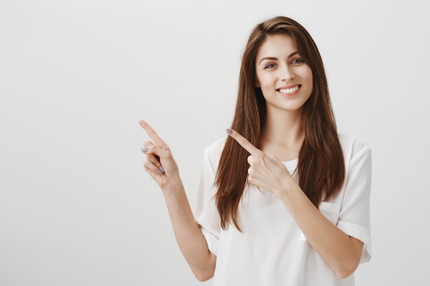 Pleased beautiful woman pointing at upper left corner, smiling happy as recommend product