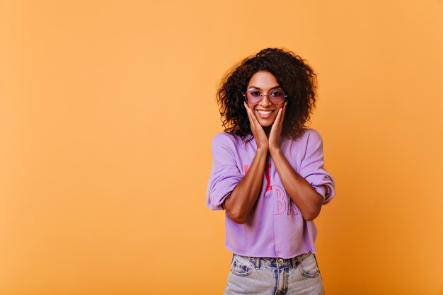 Pleased black girl in trendy sunglasses having fun during indoor shotshoot. Refined stylish african woman playfully smiling on yellow.