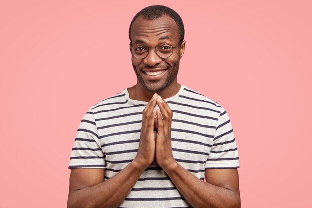 Pleased black man with satisfied expression makes praying gesture, smiles positively 