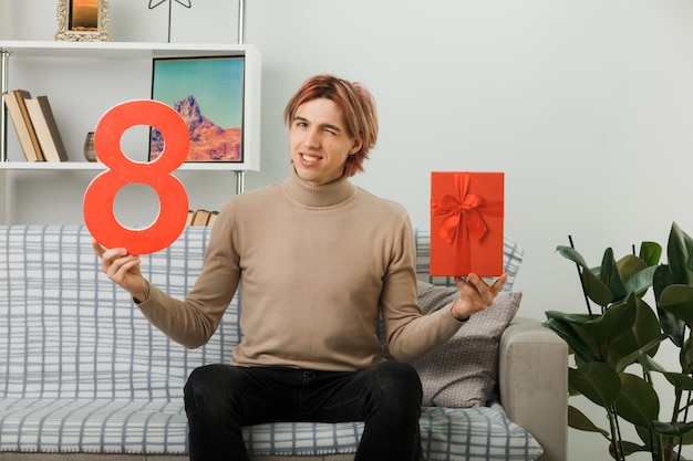 Pleased blinked handsome guy on happy women day holding number eight with present sitting on sofa in living room
