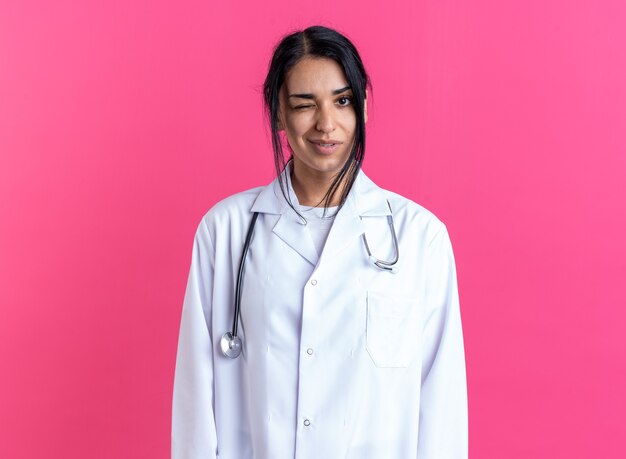 Pleased blinked young female doctor wearing medical robe with stethoscope isolated on pink wall