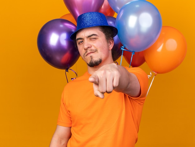 Pleased blinked young man wearing party hat standing in front balloons points at front isolated on orange wall