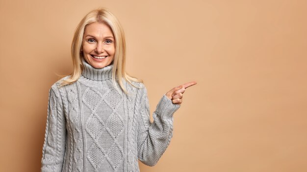 주름을 가진 기쁘게 금발 중간 나이 든된 여자는 복사 공간을 가리키는 따뜻한 회색 스웨터를 착용