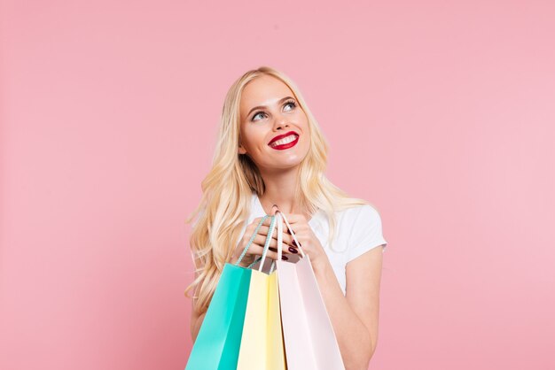 Pleased blonde woman holding packages and looking up over pink 