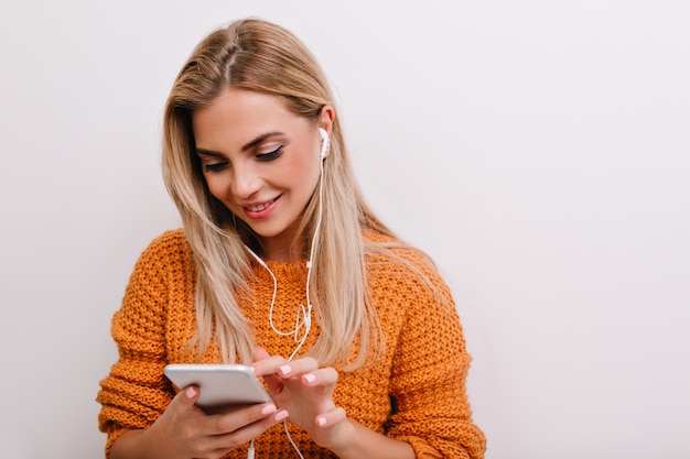 Foto gratuita donna bionda soddisfatta con trucco nudo utilizza lo smartphone mentre invia un messaggio di testo con un sorriso