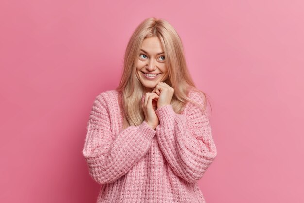 pleased blonde young woman keeps hands together and has dreamy positive face expression dressed in loose knitted sweater