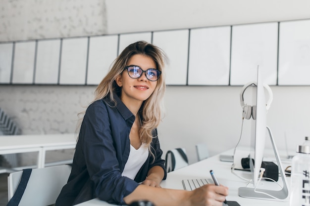 Pleased blonde young woman posing at her workplace and holding stylus