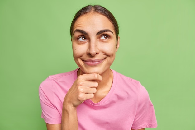 Foto gratuita una donna europea bruna contenta tiene i sogni del mento su qualcosa di concentrato sopra indossa una maglietta rosa casual isolata su un muro verde vivido