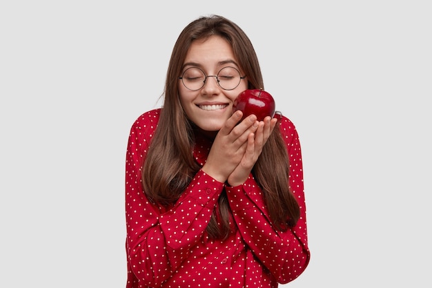 Pleased brunette woman feels pleasure, holds tasty apple, being in high spirit, wears red clothes