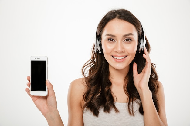 Pleased brunette woman in headphones showing blank smartphone screen and looking at the camera over gray