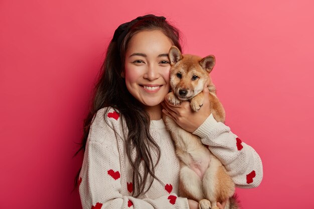 Pleased brunette woman plays with pedigree dog, embraces shiba inu, enjoys spare time, expresses loyalty from four legged friend, carries animal to vet clinic, has toothy smile.