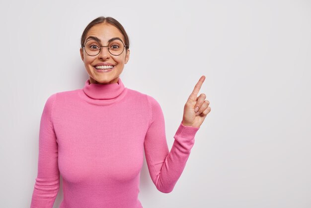 Pleased brunette woman points index finger at blank space recommends product has cheerful expression wears round spectacles casual pink turtleneck isolated over white background Look at this