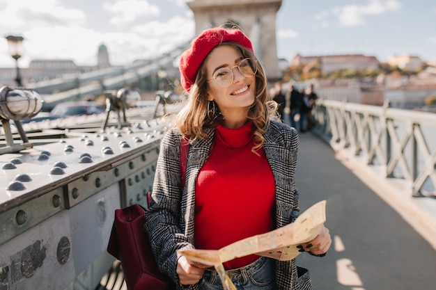 Pleased brunette woman with red backpack posing on bridge on urban background