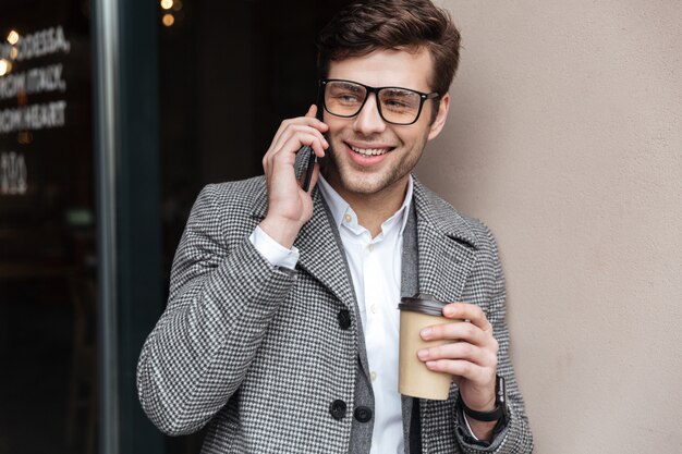 Pleased businessman in eyeglasses and coat talking by smartphone