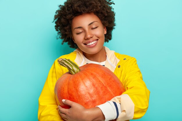 Pleased calm Afro woman harvesting pumpkin, tilts head, closes eyes and smiles broadly, wears yellow raincoat, enjoys autumn time and holidays, isolated