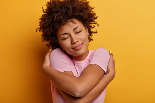 Pleased calm curly ethnic woman feels good, hugs herself